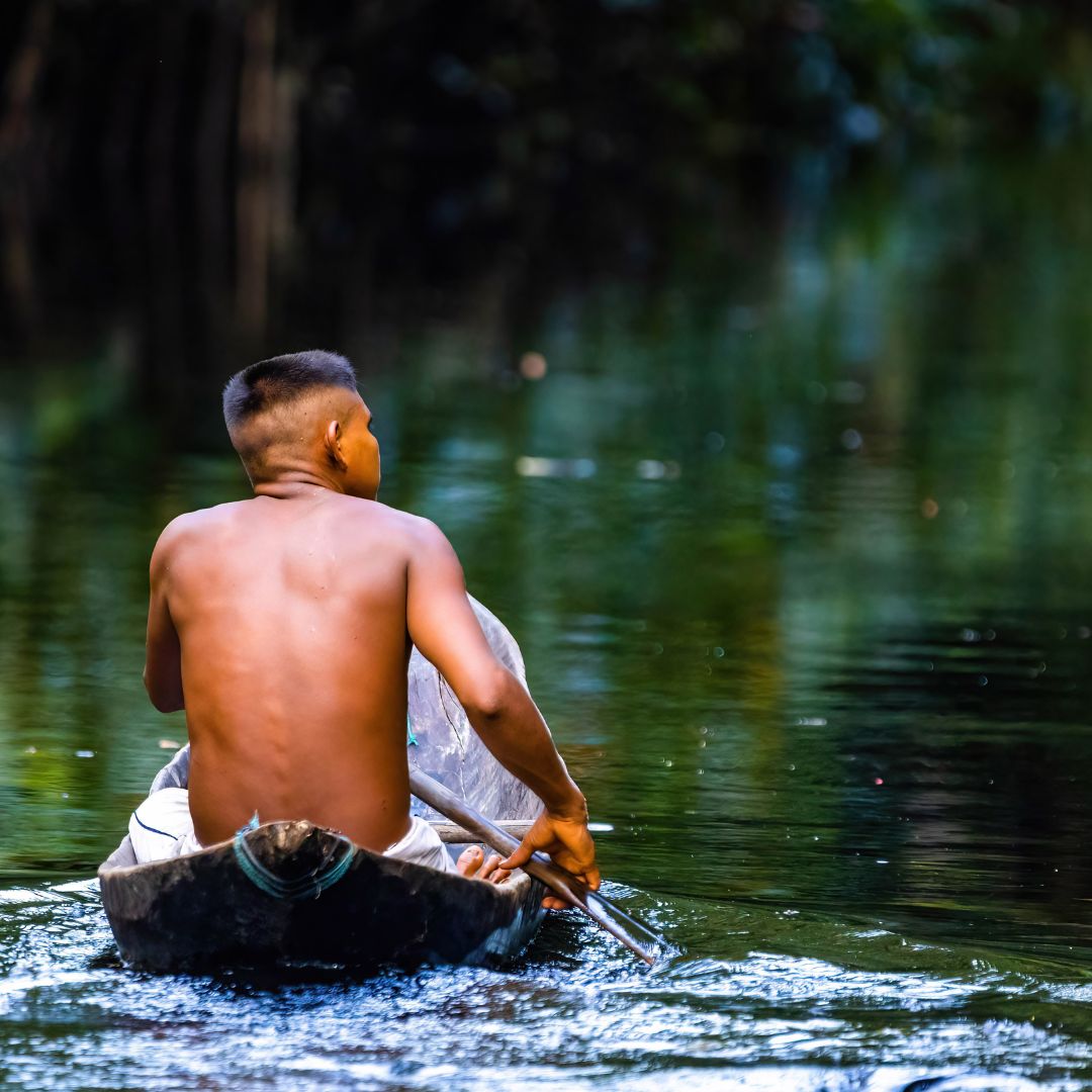 A young boy rows a small wooden boat through calm, green waters, with trees reflected in the surface. He is shirtless, focused on navigating the waterway.