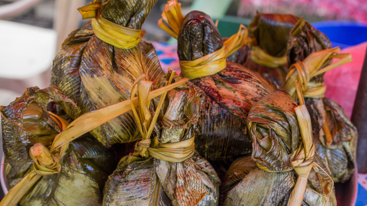 A cluster of wrapped food parcels made from bamboo leaves, tied with thin strips of palm leaves. The parcels have a glossy, dark green appearance, indicating they may contain rice or other ingredients.