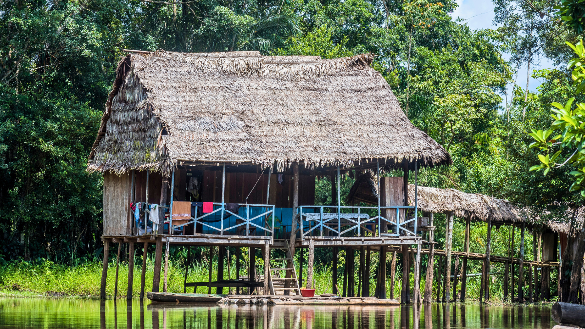 A thatched-roof hut elevated on stilts stands by a serene river, surrounded by lush greenery and trees. The structure features a balcony with colorful fabric hanging, adding vibrancy to the natural setting.