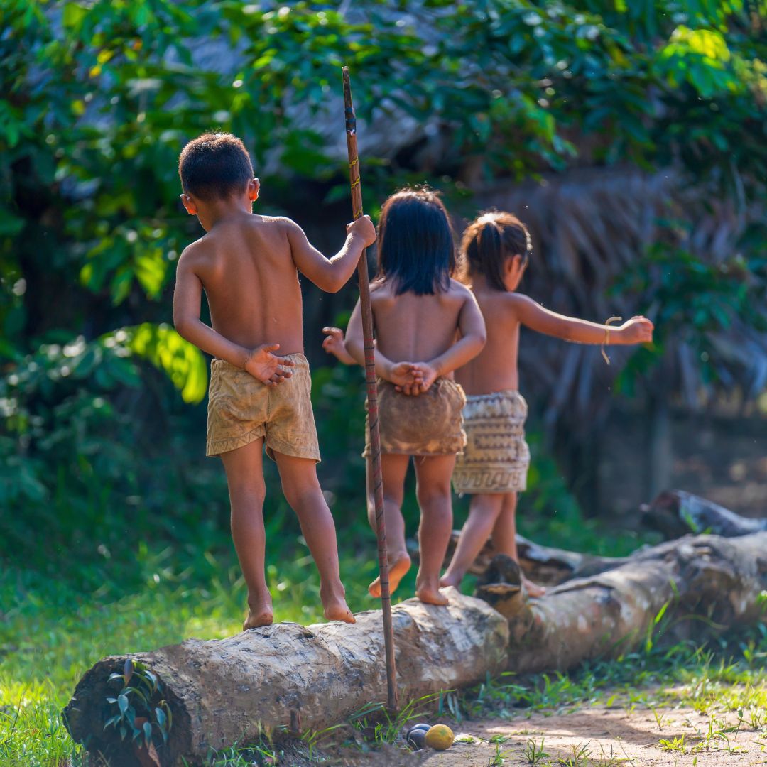 Three children stand barefoot on a log in a lush, green outdoor setting. Two boys and a girl are dressed in simple shorts, with the girl holding a small stick. They appear playful, with hands behind their backs, surrounded by dense foliage.