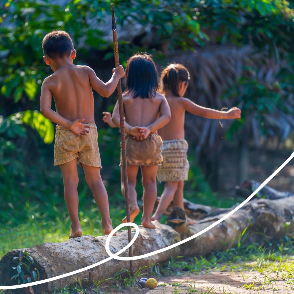 Three children play on a fallen log in a lush, green setting. The children are mostly bare, with one holding a stick, and they appear to be balancing as they walk along the log. Their backs are turned towards the viewer, showcasing a natural, carefree moment.
