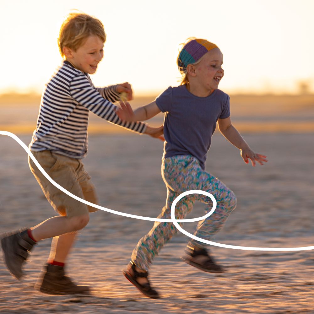 Two joyful children running together on a sandy beach at sunset. The boy, wearing a striped shirt and shorts, holds hands with the girl, who has colorful leggings and a cap, both laughing and enjoying the moment.