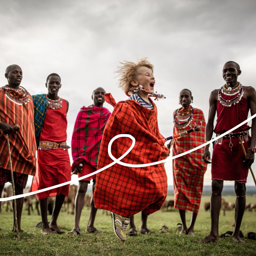 A young child joyfully jumps in front of a group of Maasai men dressed in traditional attire, standing together in a grassy landscape under a cloudy sky.