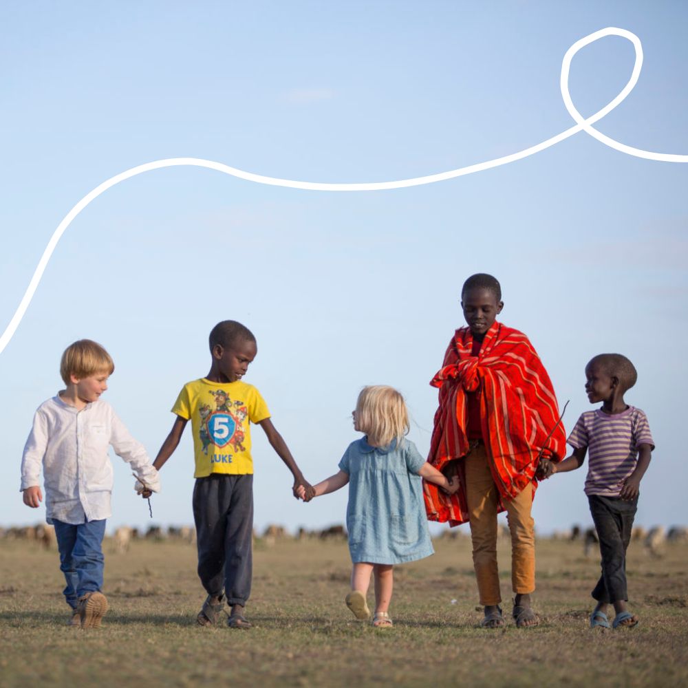 A group of five children, including a boy in a white shirt, two boys in casual tops, a girl in a blue dress, and a boy wearing a red and white striped shirt, hold hands while walking on a grassy field. They are smiling and enjoying each other's company against a clear blue sky.