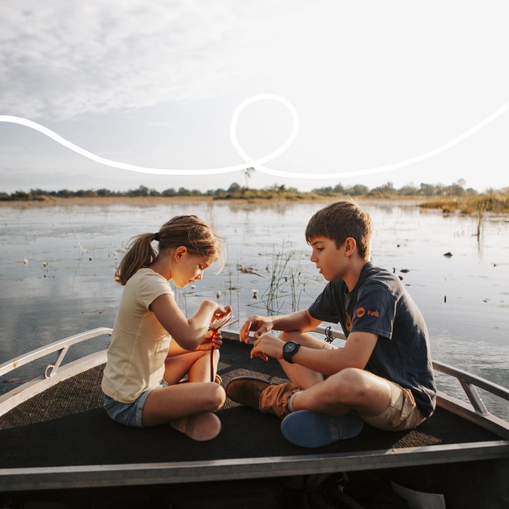 Two children sitting on the edge of a boat, engaged in an activity. One child is tying something with a string, while the other observes. The background features a serene water landscape with vegetation and a cloudy sky.
