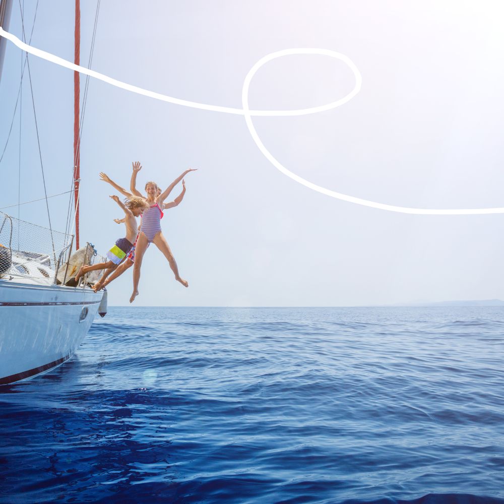 Two children joyfully jumping off a sailboat into the ocean on a sunny day. The water is calm and blue, with a bright, clear sky in the background.