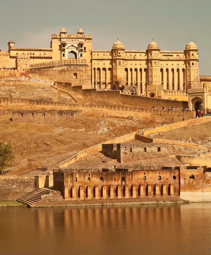 Historic Amber Fort in Rajasthan, India, showcases stunning golden architecture on a hillside, reflecting the rich cultural heritage. A serene body of water at the base enhances the majestic view of the fort's intricate structures and stairs.