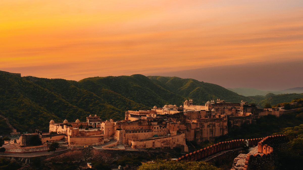 A majestic fort located on a hillside, surrounded by lush green mountains, with a vibrant sunset sky in the background. The fort features a blend of architectural styles and is encompassed by ancient walls.