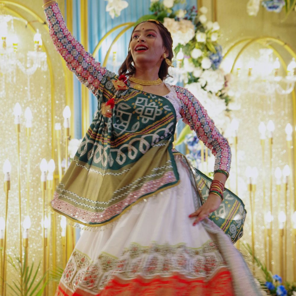 A woman gracefully dances in a colorful traditional outfit with intricate patterns, featuring a green and white top and a flowing, embroidered skirt. She has joyful expression and traditional jewelry, with decorative lights and floral arrangements in the background.