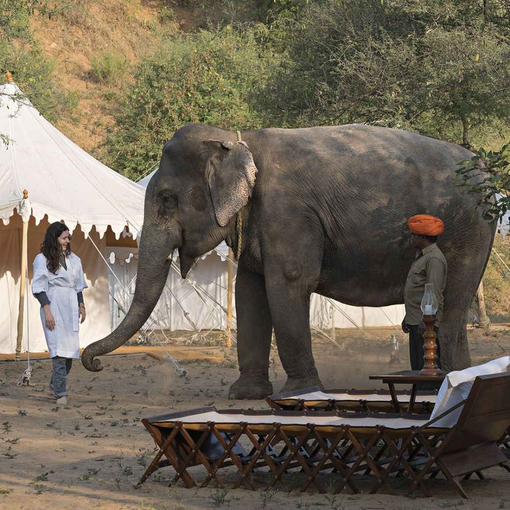 A woman in a white outfit interacts with a large elephant in a desert-like setting. Nearby, a man in traditional attire observes, with tents and wooden furniture in the background.