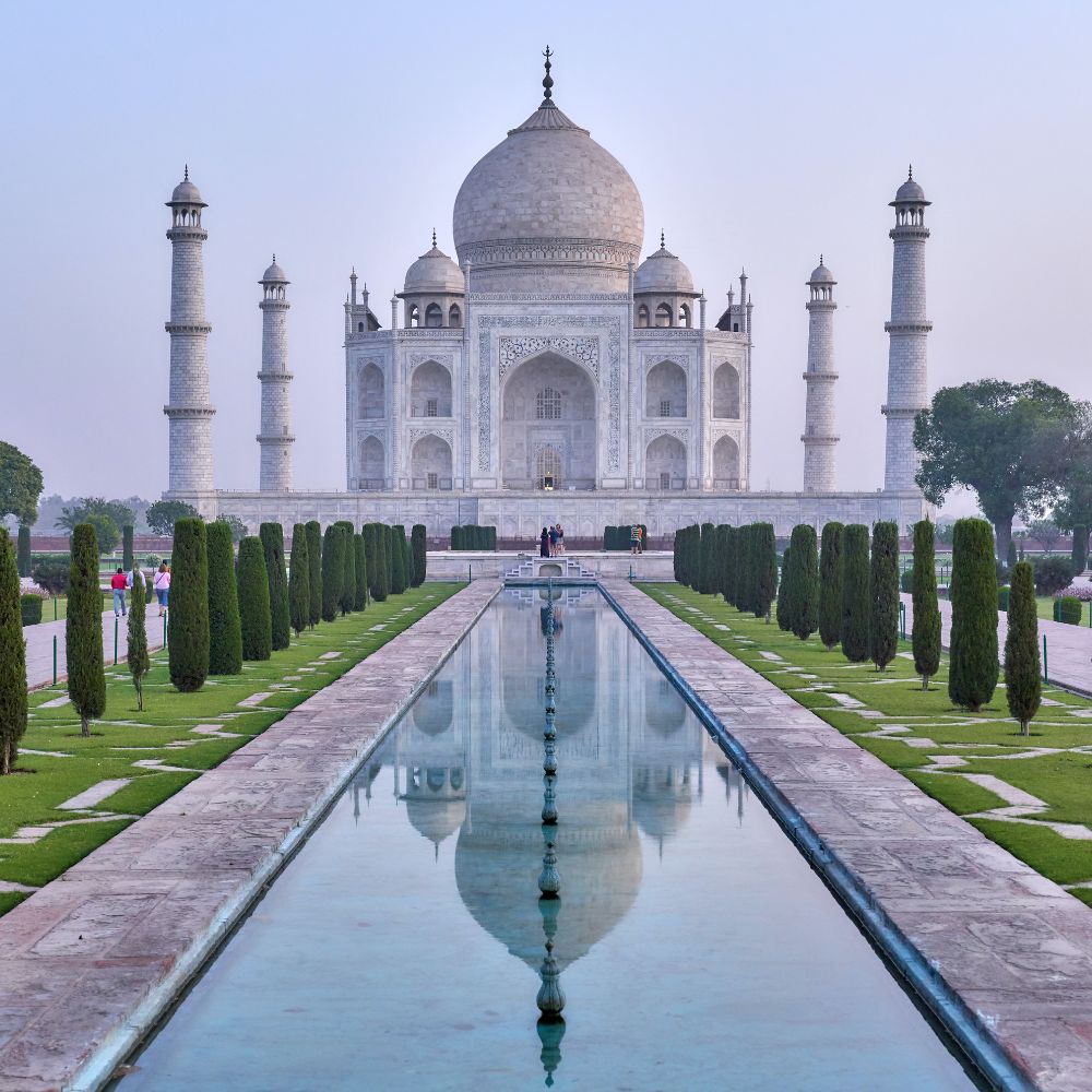 The Taj Mahal stands majestically with its white marble dome and minarets, reflecting in a serene water channel. Lush green gardens and neatly trimmed trees line the pathway, creating a picturesque scene.