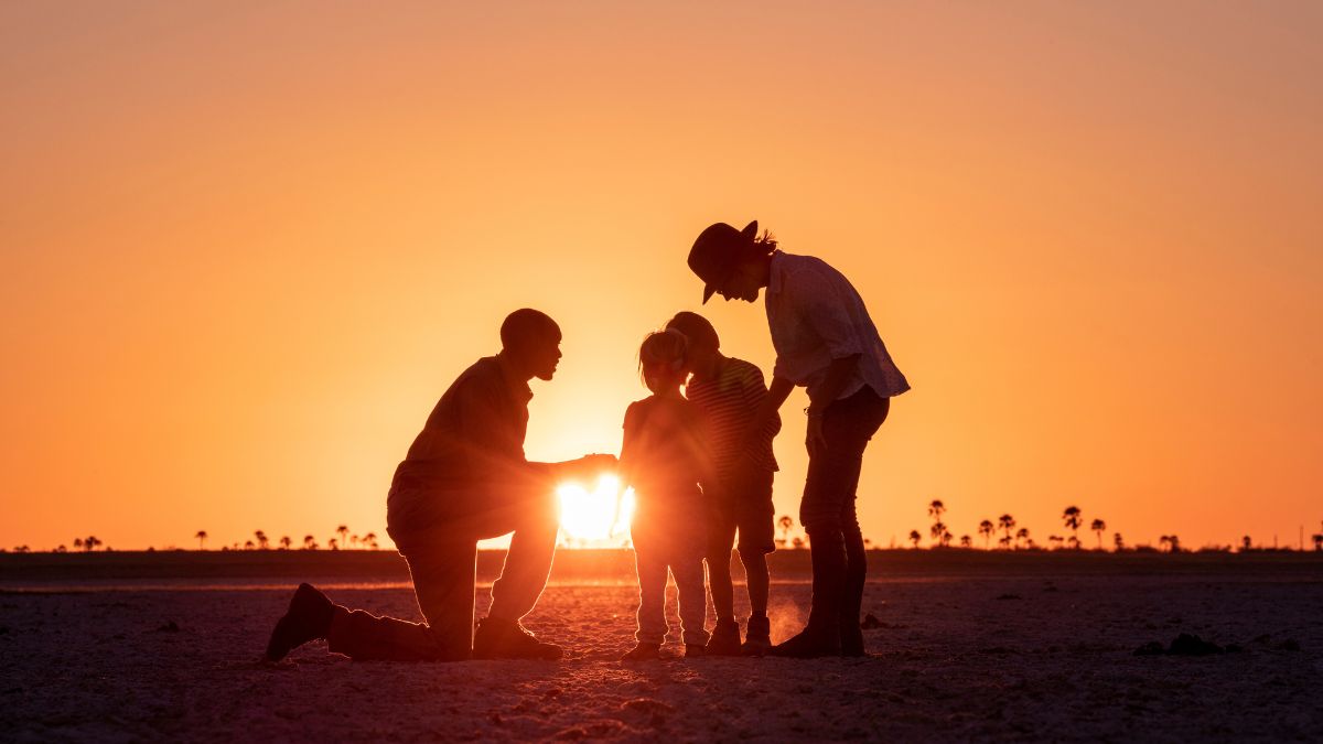 A silhouette of a family at sunset, with two adults kneeling beside two children. The sun sets behind them, casting a warm glow over the scene and palm trees in the distance.