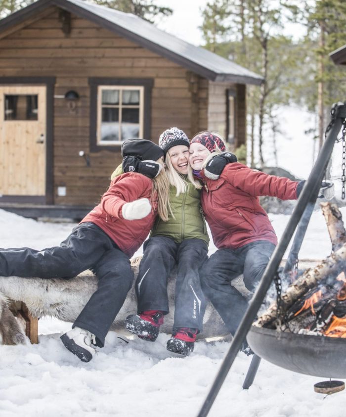Three children sit on a snowy bench, smiling joyfully while wearing winter jackets and hats. A warm fire burns nearby, and a cozy wooden cabin is visible in the background.