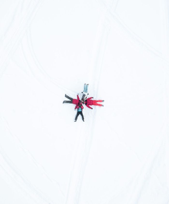 Five people in brightly coloured winter clothes lie on a snow-covered surface, arranged in a star shape with their arms and legs extended. The surrounding snow is untouched, creating a stark contrast with their vibrant attire.