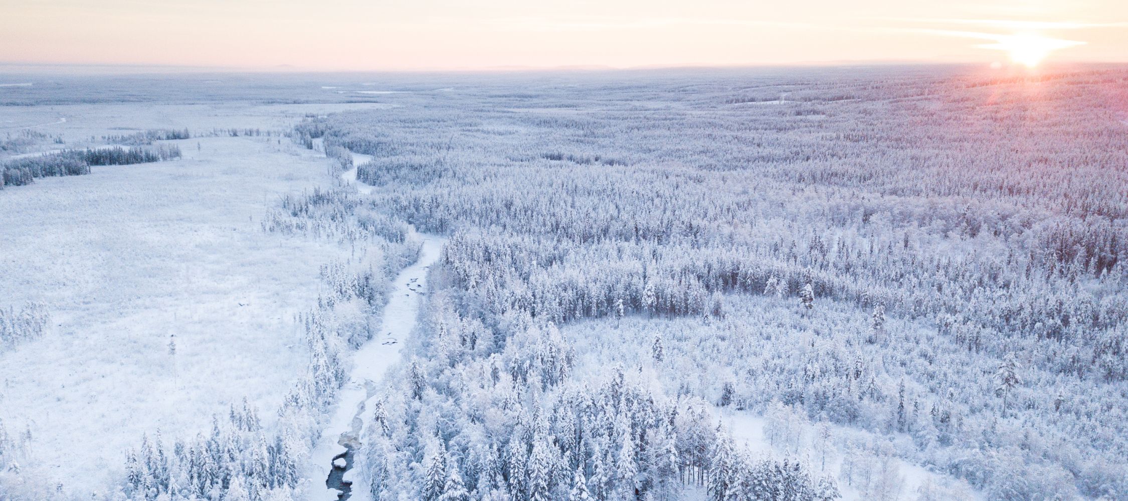 A breathtaking aerial view of a snow-covered landscape with a meandering river cutting through dense forests, illuminated by a soft sunrise in the distance.