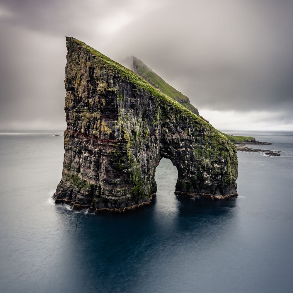 A towering sea stack rises from calm waters, featuring a large arch on one side and lush green vegetation on top. Gray clouds loom overhead, creating a dramatic atmosphere.