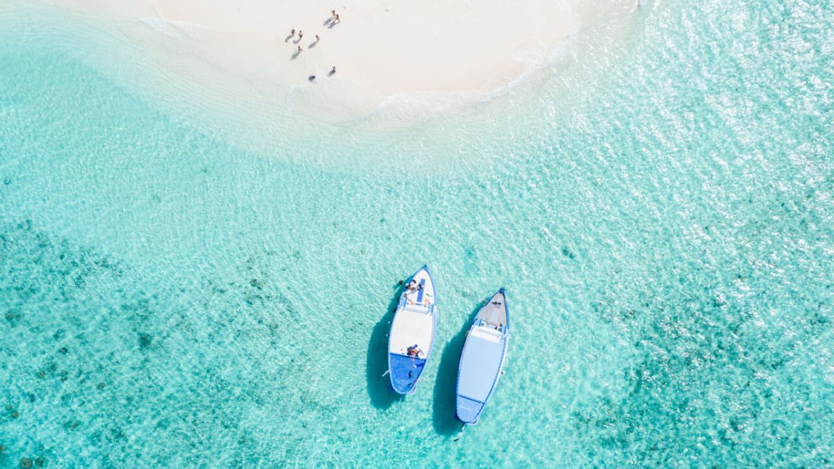 Two boats are anchored in clear turquoise water near a sandy beach. A few people are lounging on the shore, enjoying the sunny day.