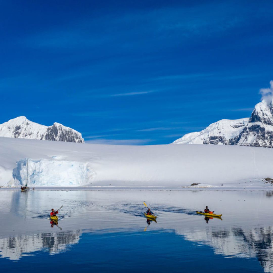 Three kayakers paddling on a calm, reflective body of water surrounded by snow-covered mountains and bright blue skies. Glaciers can be seen in the background, adding to the stunning icy landscape.
