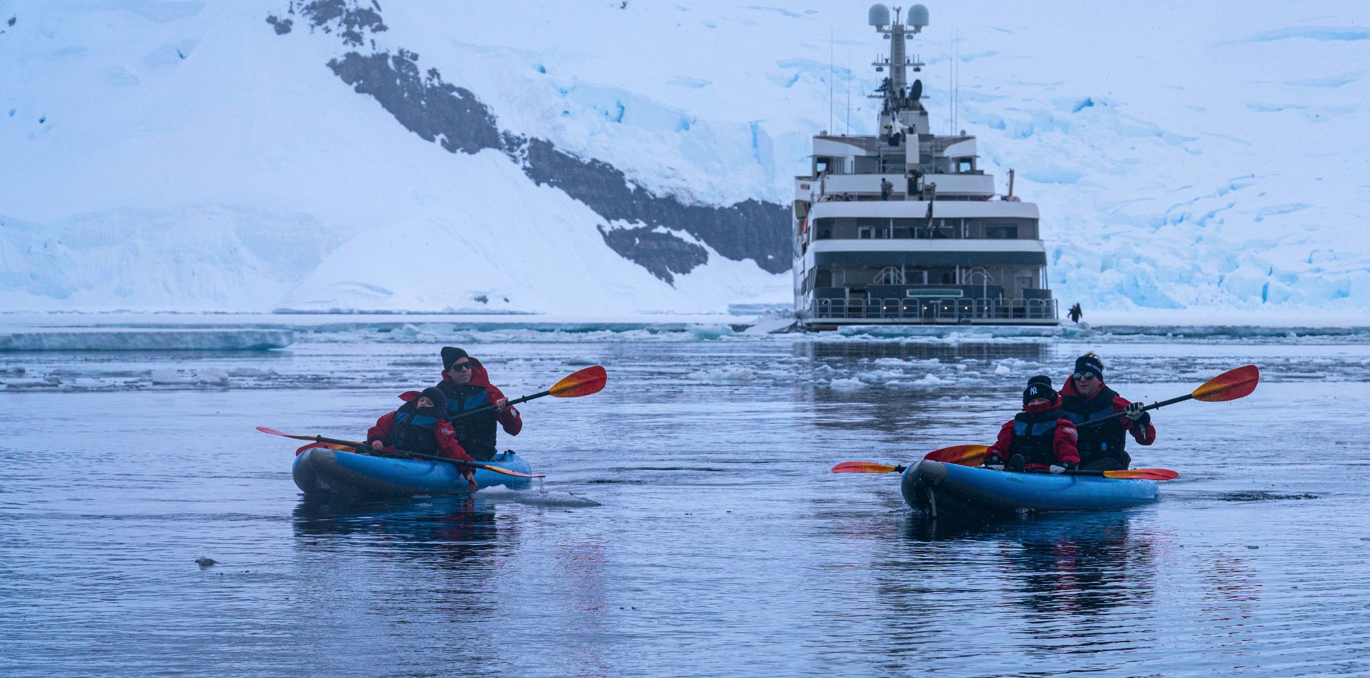 Two kayakers in red jackets and life vests paddle in icy waters, with a large ship anchored in the background amidst snow-covered cliffs and glaciers.