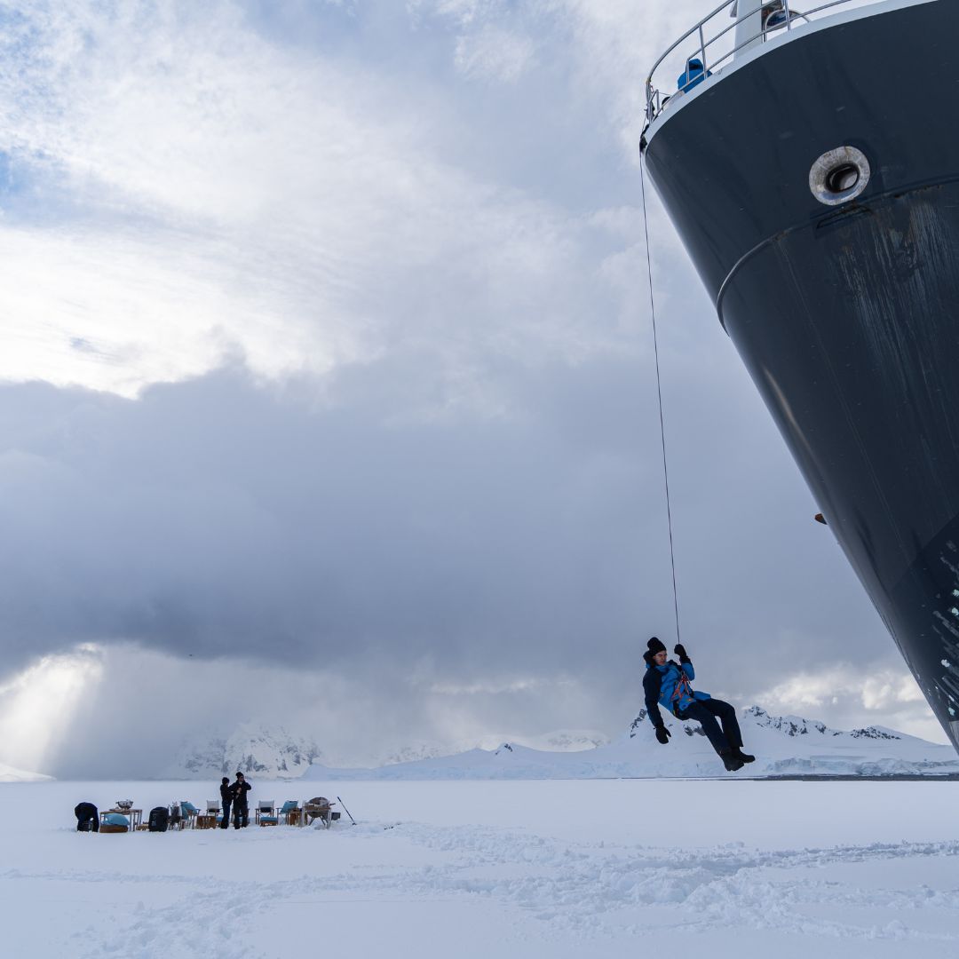 A person is being lowered from a ship on a rope onto a snowy landscape, with a group of people working in the background amidst mountains and cloudy skies.