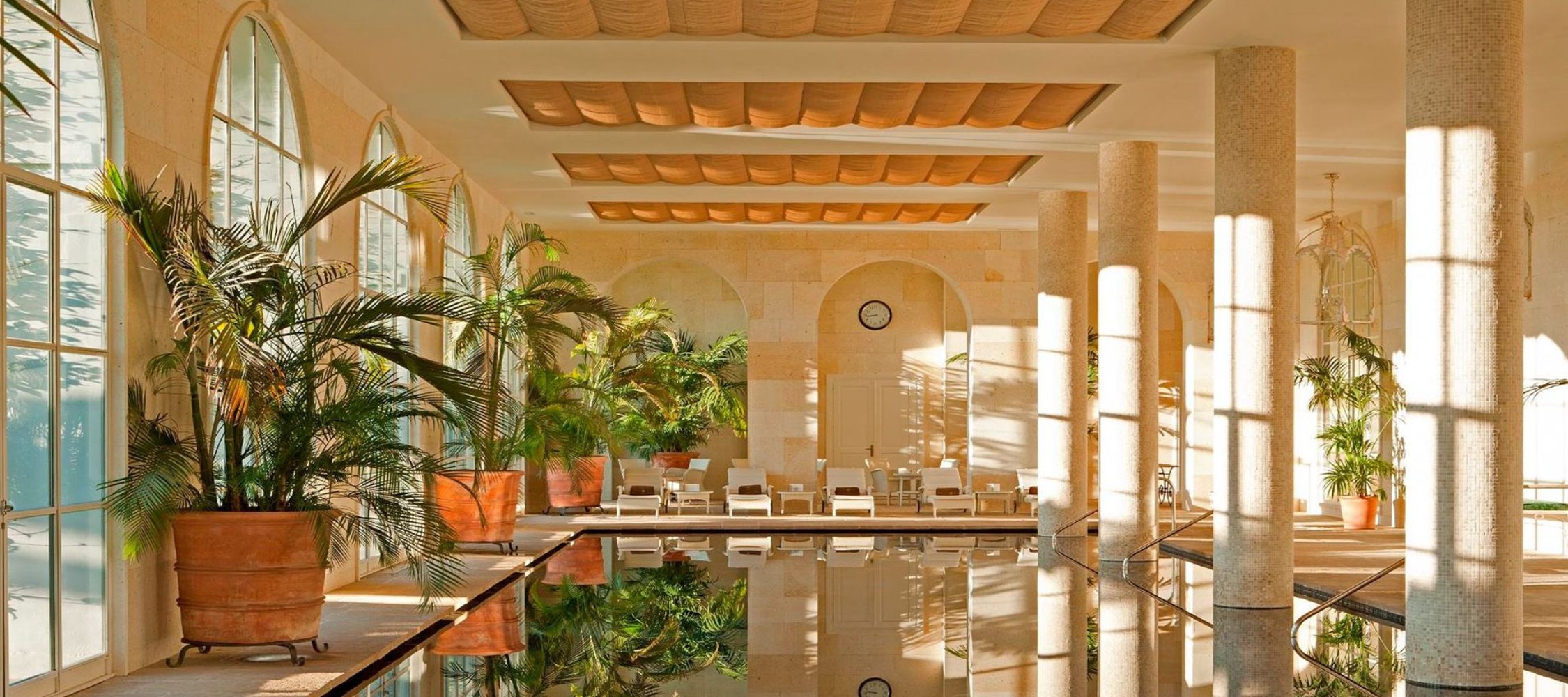 A serene indoor pool area with large windows allowing natural light, surrounded by tall columns and lush greenery. Comfortable loungers are positioned around the pool, and a clock is mounted on the wall. The atmosphere is calm and elegant.