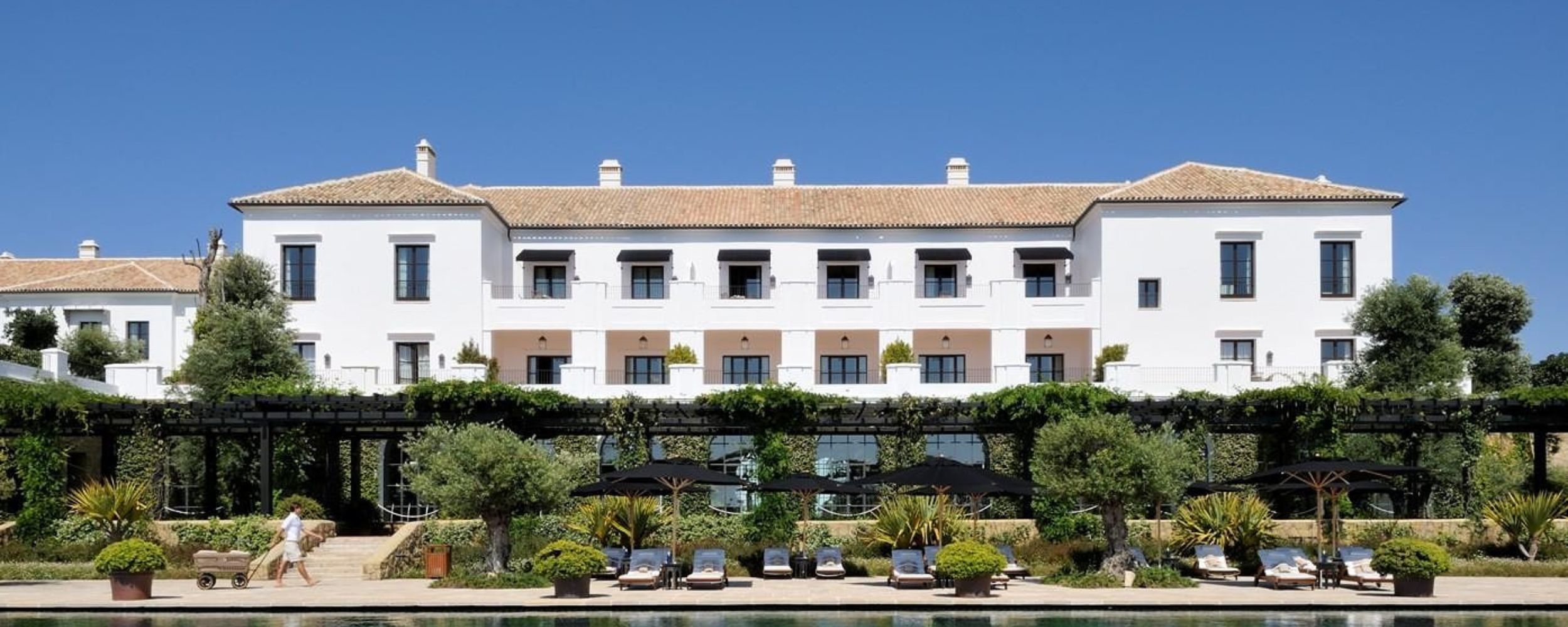 A luxurious white hotel with a terracotta roof surrounded by greenery, featuring a row of sun loungers and umbrellas by a reflective pool. Bright blue sky above enhances the serene atmosphere.