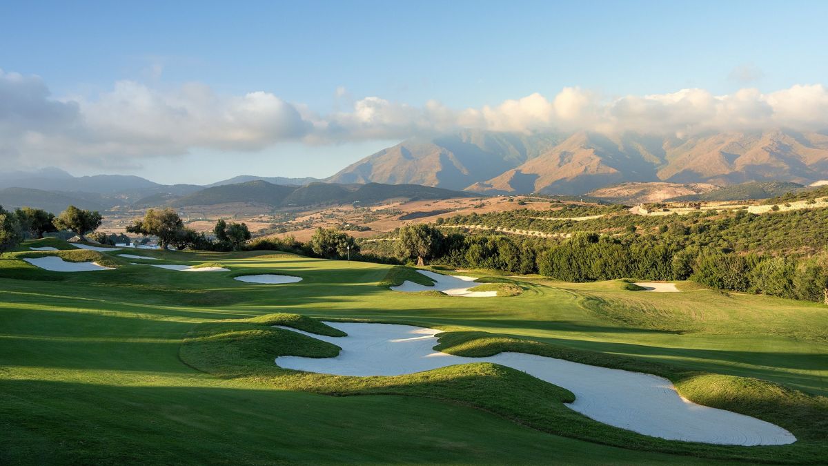 A scenic golf course with lush green fairways and white sand traps, set against a backdrop of mountains and a clear blue sky.