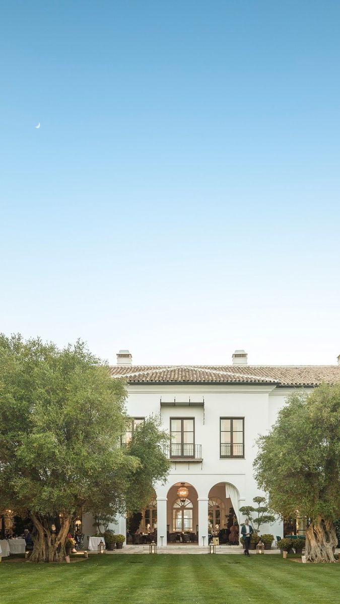 A grand white villa with arched doorways, framed by lush green trees and a clear blue sky, showcasing a serene outdoor setting.