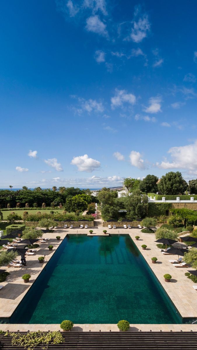 A serene outdoor scene featuring a large swimming pool surrounded by neatly manicured gardens. Lush greenery and ornamental shrubs frame the pool, with sun loungers and umbrellas positioned around it. A clear blue sky with scattered clouds complements the tranquil atmosphere.