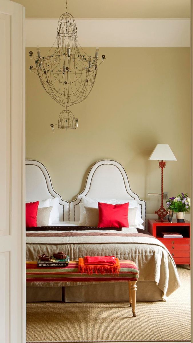 A stylish bedroom with a double bed featuring a light-coloured headboard and red cushions. A striped bench at the foot of the bed is adorned with a bright throw. A chandelier made of wire hangs from the ceiling, and a lamp stands on a red bedside table beside the bed. The walls are a soft green, creating a warm atmosphere.