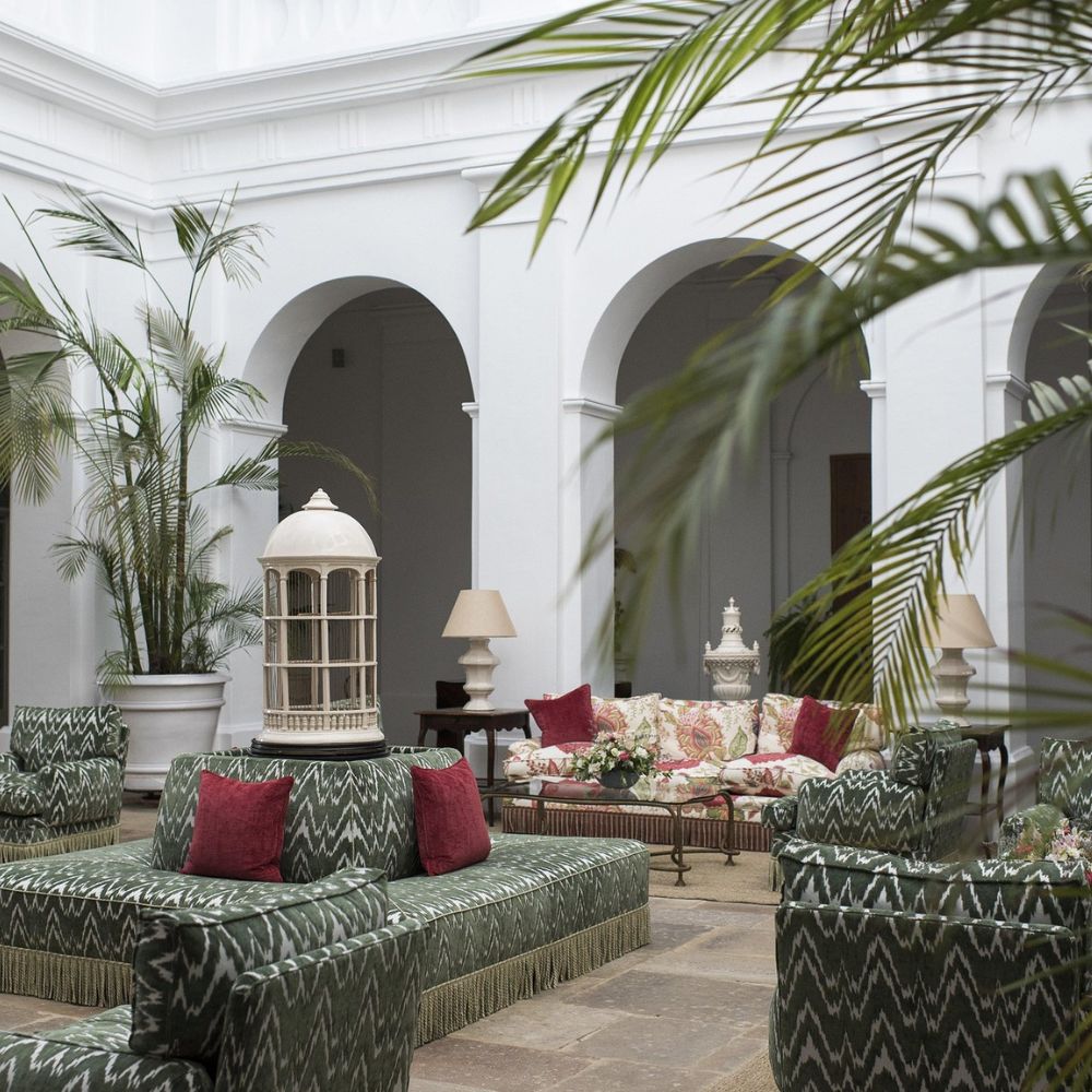 A stylish indoor space featuring lush green plants, patterned seating, and elegant furniture. The room has high white walls and arched doorways, creating an airy atmosphere. The decor includes a decorative lantern and floral cushions.