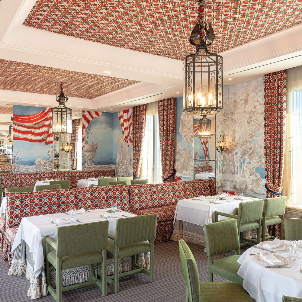A stylish restaurant interior featuring patterned red and white wallpaper and ceiling, green upholstered chairs, and elegantly set tables with white tablecloths. Decorative lanterns hang from the ceiling, and large windows allow natural light to fill the space.