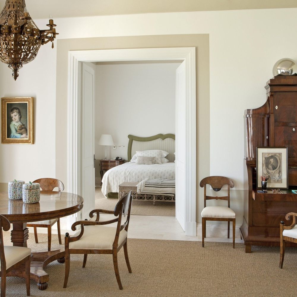 A spacious, elegantly decorated room features a wooden dining table with chairs, a vintage writing desk, and a doorway leading to a cozy bedroom with a large bed and soft lighting. A chandelier hangs from the ceiling, adding a touch of sophistication.