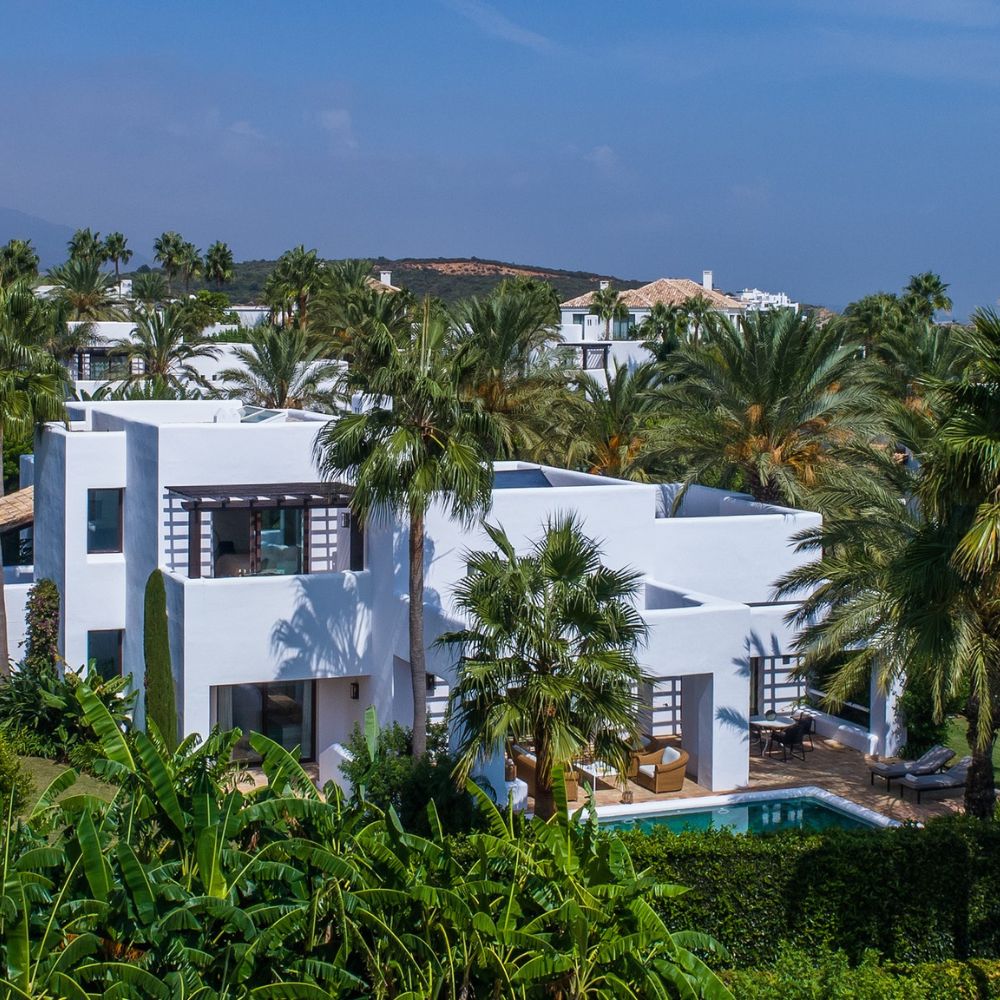 Modern white villa surrounded by palm trees and lush greenery, featuring a pool and a tranquil outdoor seating area. Mountains loom in the background under a clear blue sky.