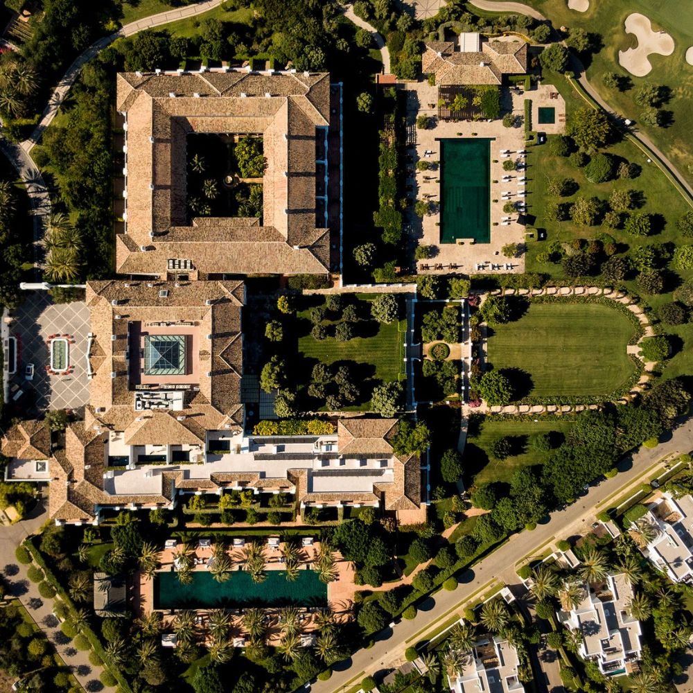 Aerial view of a luxurious resort complex featuring several buildings with terracotta roofs, surrounded by landscaped gardens, swimming pools, and manicured lawns. Nearby are tennis courts and a golf course, showcasing a lush, green environment.