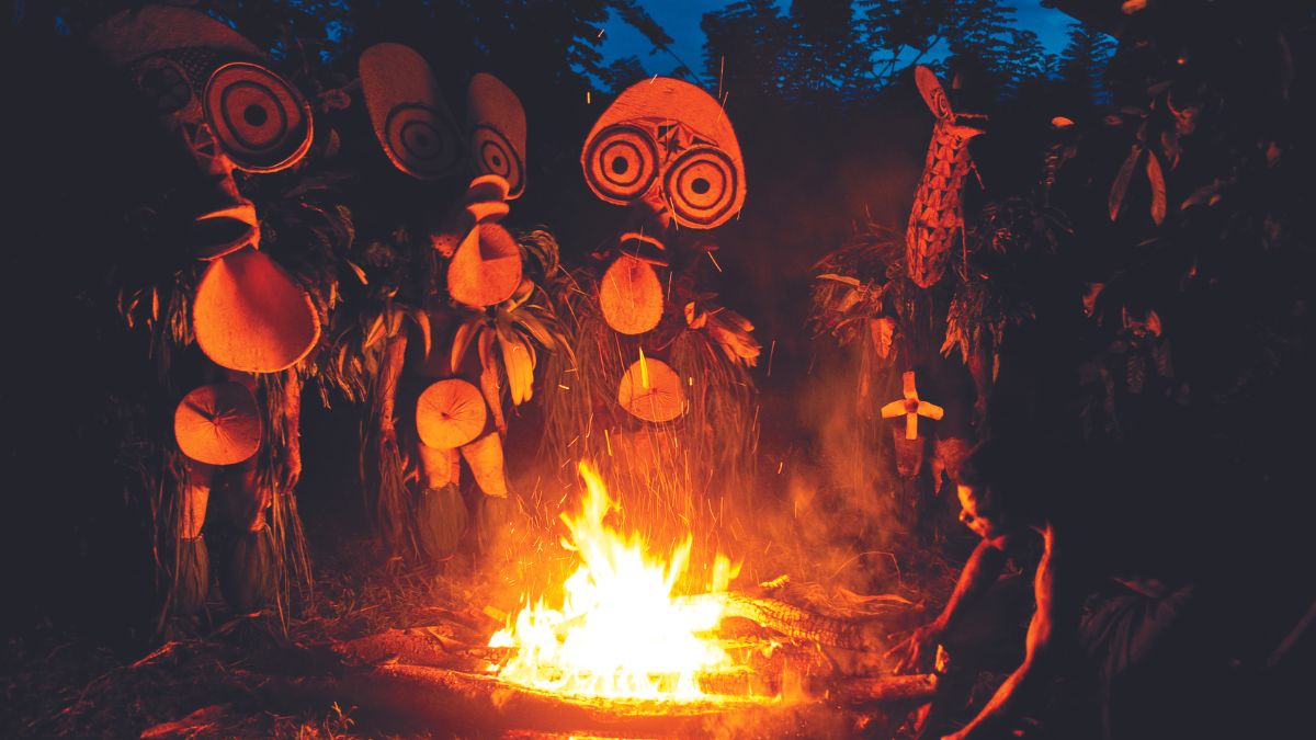 A group of individuals in traditional attire with elaborate masks stands around a large fire in a forested area, creating a mystical atmosphere. The scene is illuminated by the firelight, highlighting their costumes and the surrounding vegetation.
