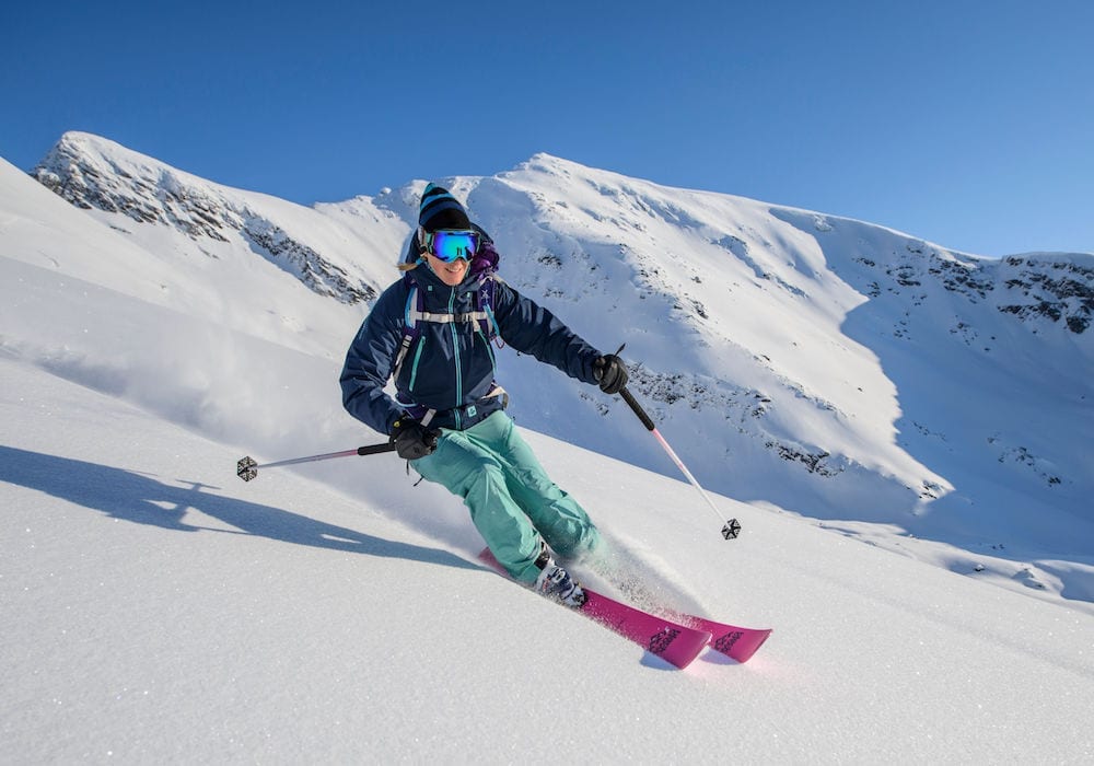 A skier navigates down a snowy slope, wearing a blue jacket, teal pants, and purple skis. Snow is being kicked up behind them against a backdrop of snowy mountains and a clear blue sky.
