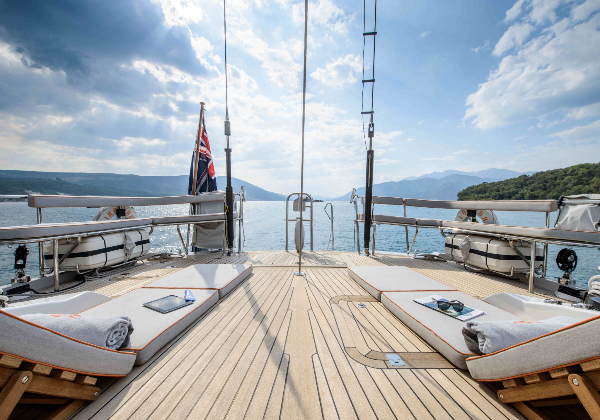 A serene view from the deck of a yacht, featuring two loungers with towels, overlooking calm waters and distant mountains under a partly cloudy sky. A flag is visible, adding to the nautical ambiance.