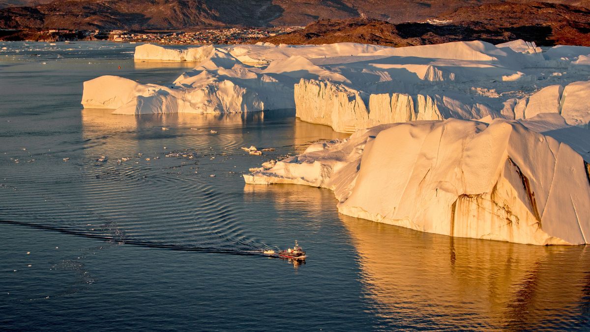 A tranquil scene of a boat navigating through icy waters near towering white icebergs, reflecting the warm light of a setting sun. The distant town and rugged mountains are visible in the background.