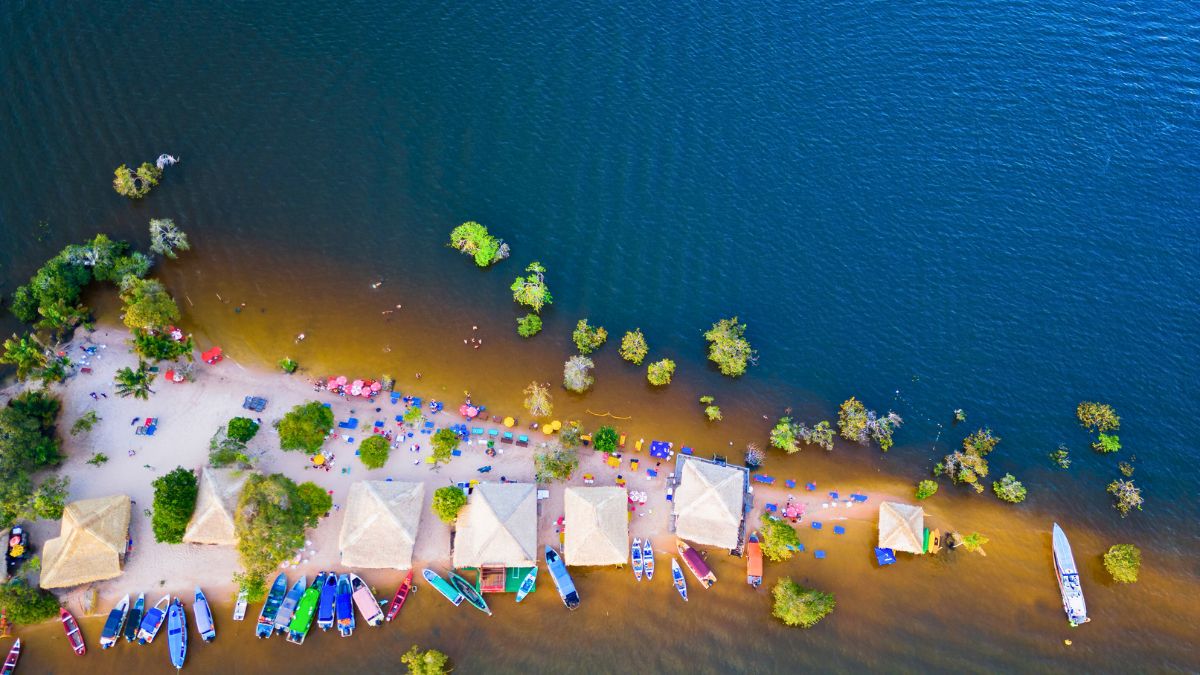 Aerial view of a sandy beach lined with thatched-roof huts and moored boats. Clear blue water surrounds the area, with people sunbathing and trees scattered along the shoreline. Brightly colored umbrellas and loungers add to the vibrant atmosphere.