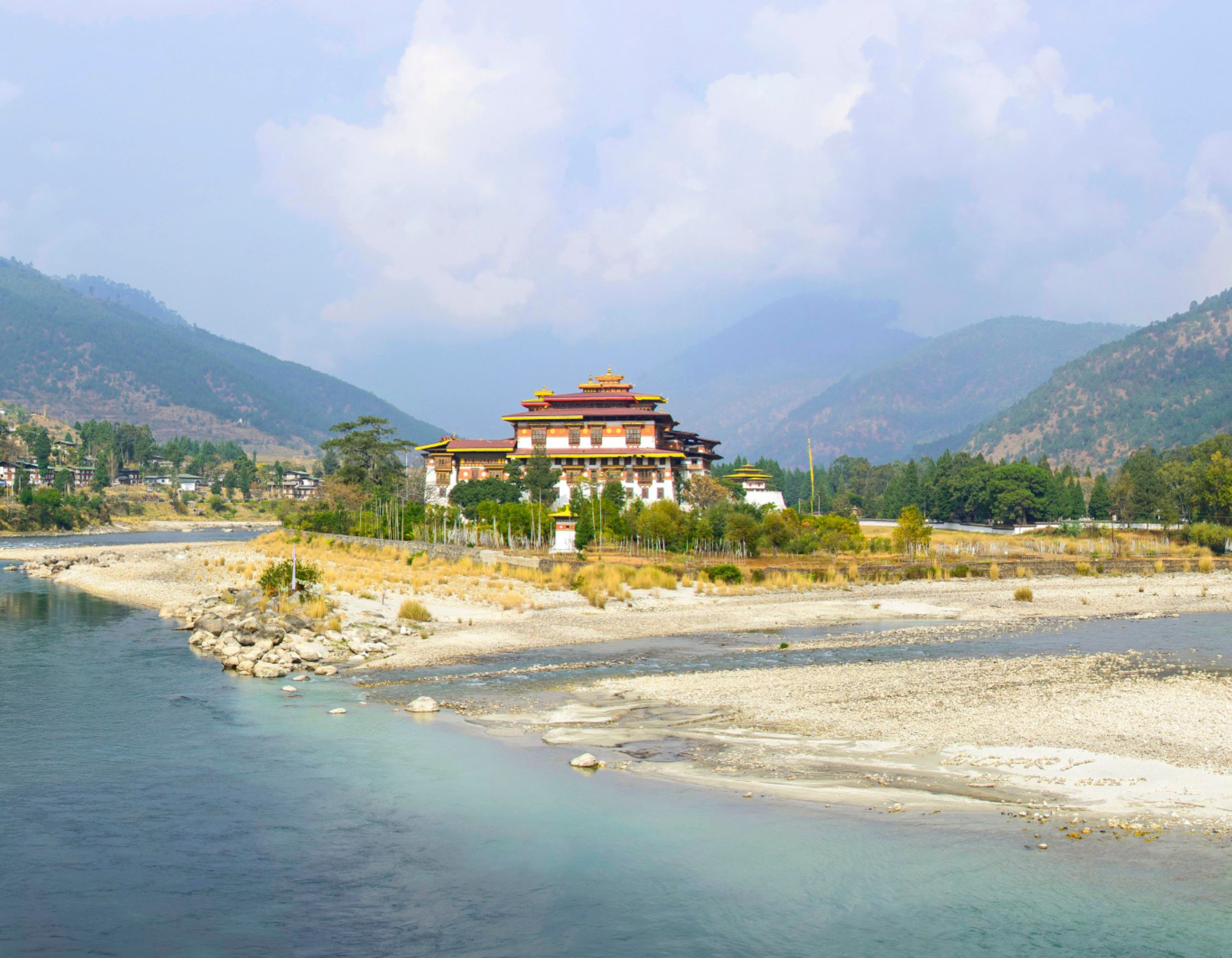 A majestic monastery nestled along a riverbank, surrounded by mountains and lush greenery. The structure features vibrant yellow and red roofs, with a serene landscape of rocky shores and a clear blue sky.