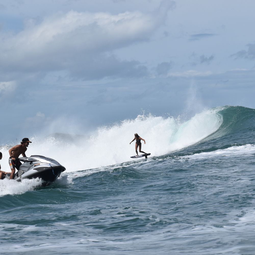 A person is surfing a large wave while being towed by a jet ski. The sky is partly cloudy, and the ocean is choppy with white foam cascading off the wave.