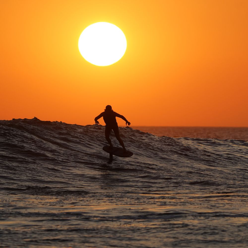Silhouetted surfer riding a wave during a vibrant sunset, with a large sun glowing on the horizon.