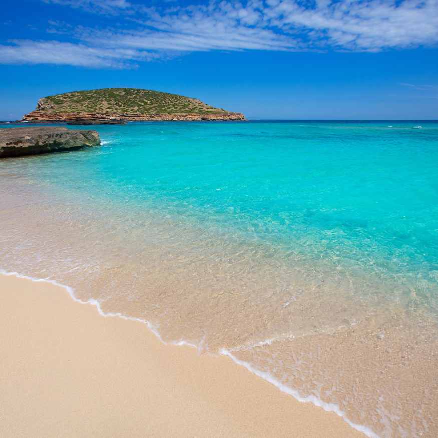 A serene beach with soft, golden sand leading to clear turquoise waters. In the background, a lush green island is visible under a bright blue sky with scattered clouds.