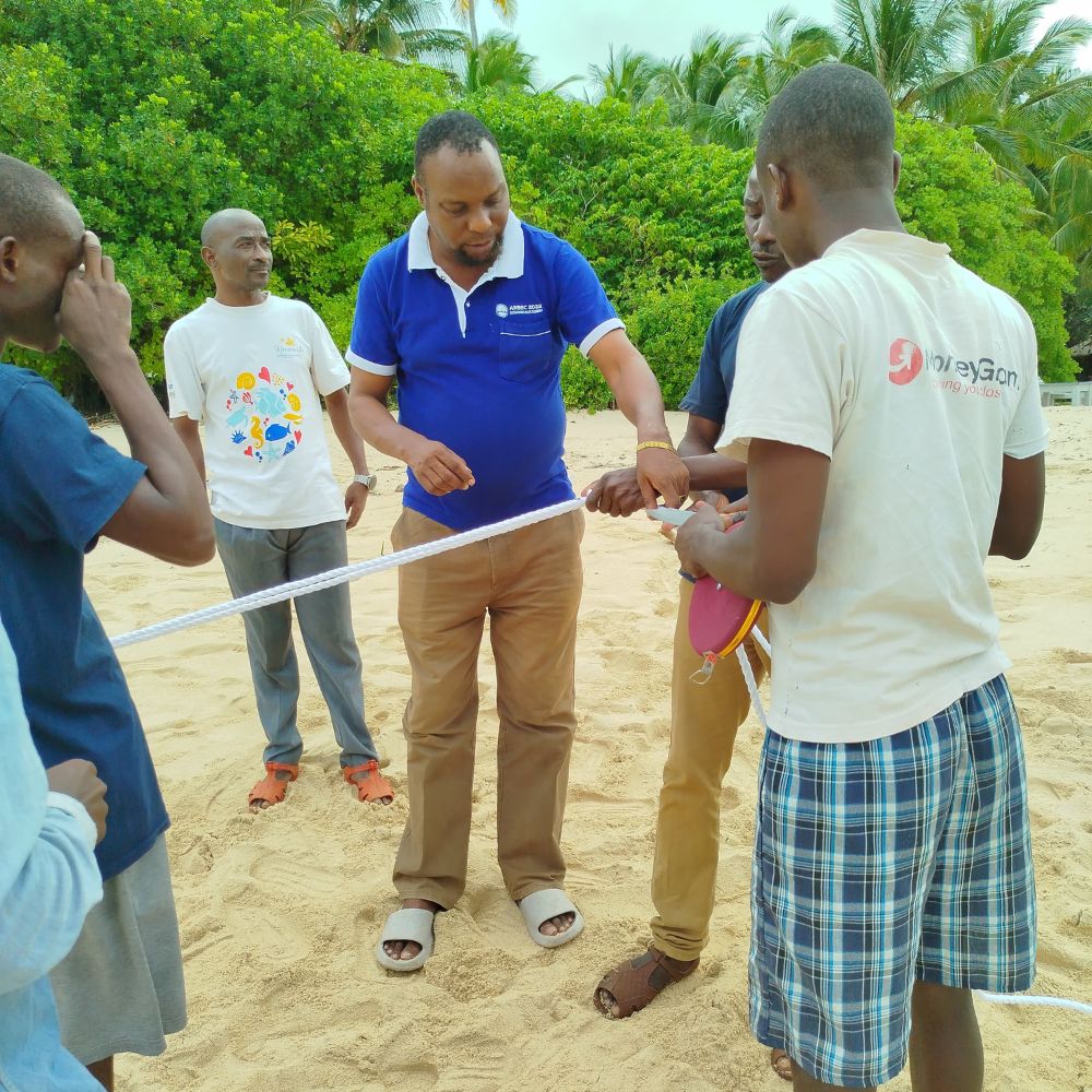 A group of men on a sandy beach are engaged in a team activity, focused on tying or measuring ropes. Some individuals are wearing casual clothing, and palm trees are visible in the background.