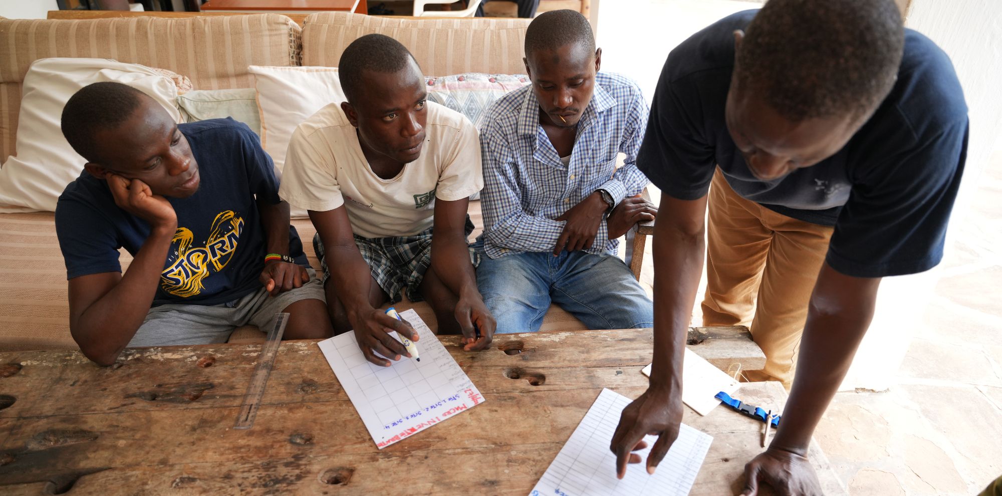 Four young men gather around a table, focused on a discussion. One man stands, pointing at notes on a sheet of paper, while the others sit attentively, with expressions of contemplation. The setting is casual, with a sofa visible in the background.