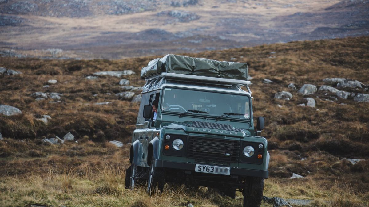 A green Land Rover Defender parked on rugged terrain, with a rooftop tent and mountains in the background. The landscape features dry grass and rocky outcrops.