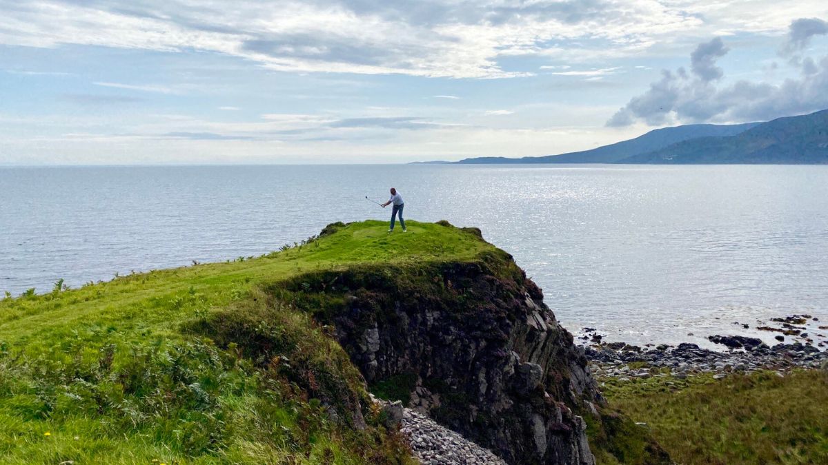 A person stands on a grassy cliff overlooking a calm sea, with rolling hills visible in the background under a partly cloudy sky.