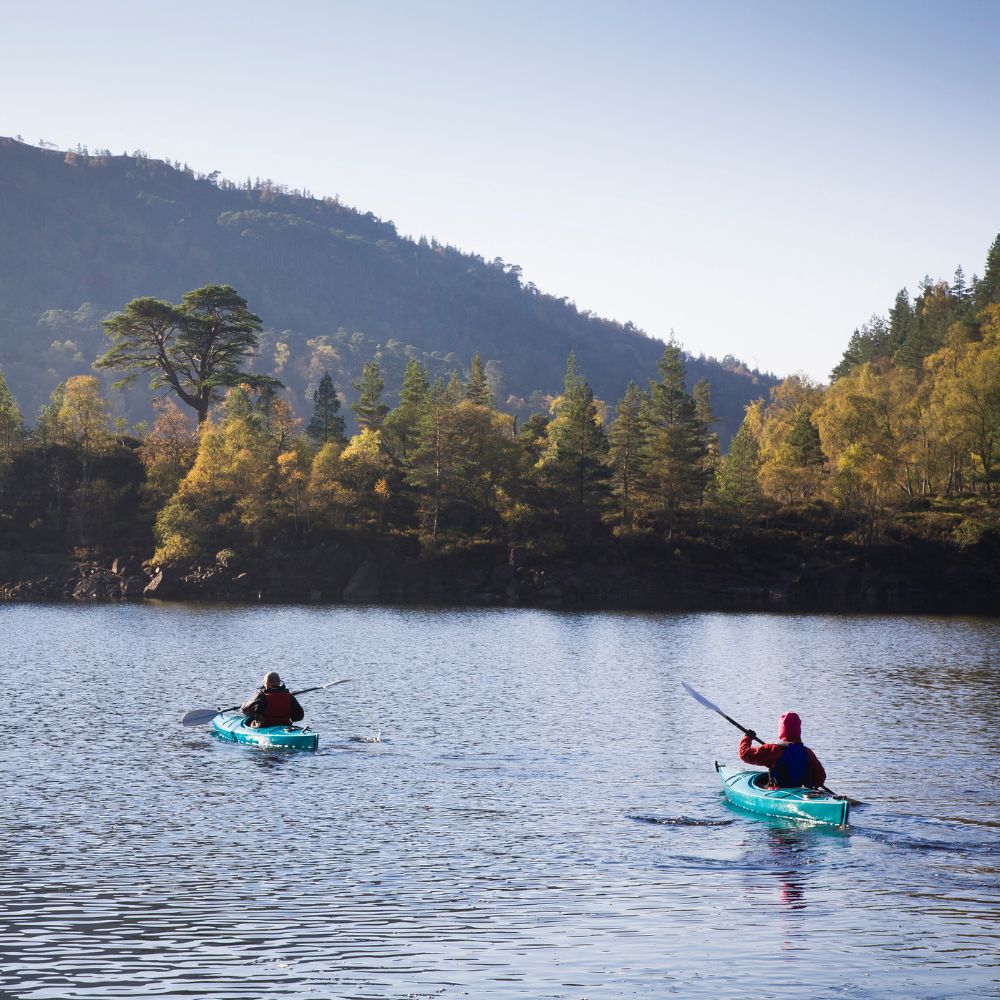 Two kayakers paddle across a calm lake surrounded by trees and mountains, with vibrant autumn foliage reflecting in the water.
