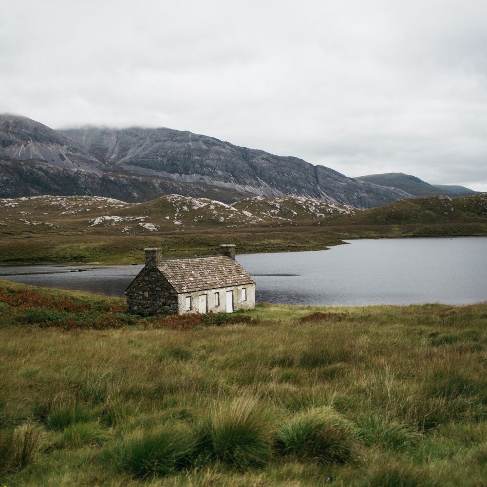 A rustic stone cottage sits next to a tranquil lake, surrounded by lush green grass and hills, with dramatic mountains in the background under a cloudy sky.